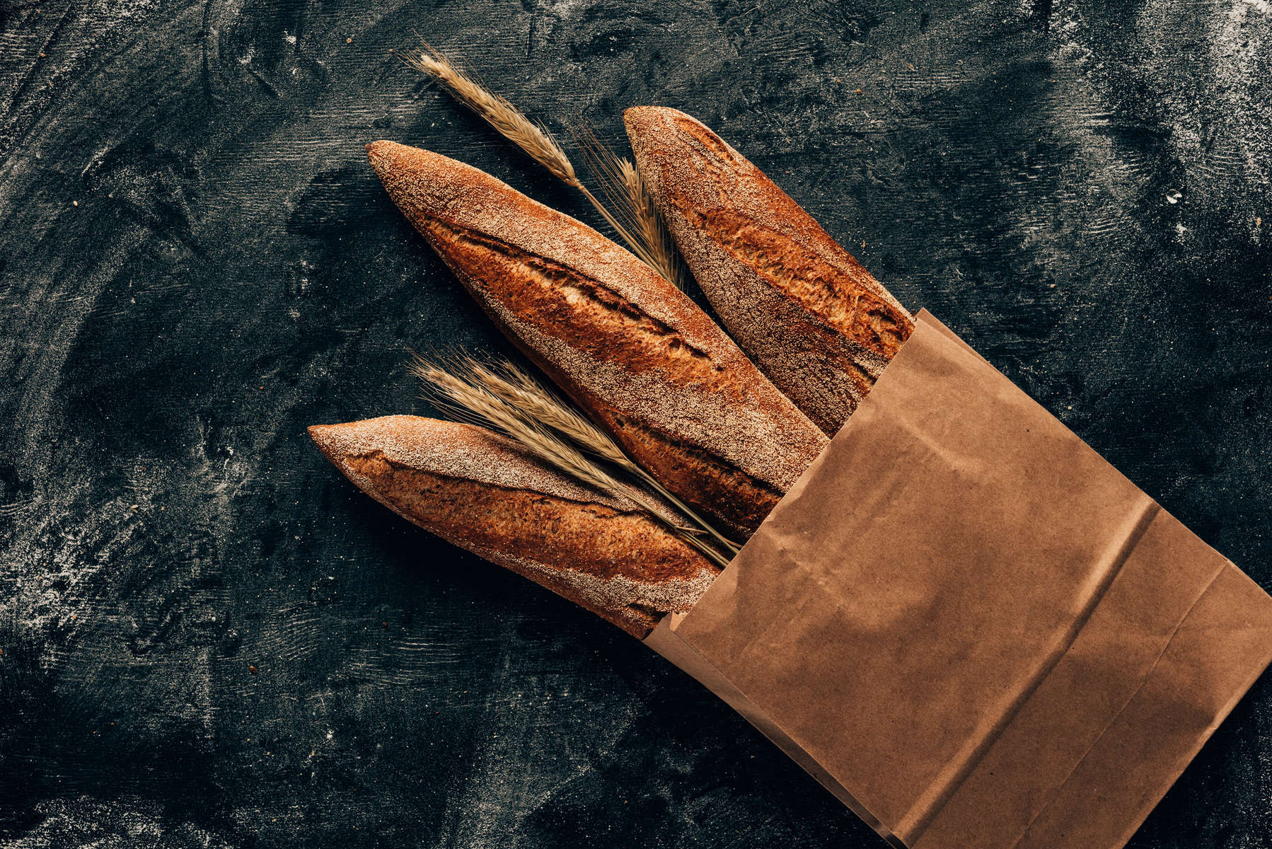 top view of arranged french baguettes in paper bag and wheat on dark tabletop with flour